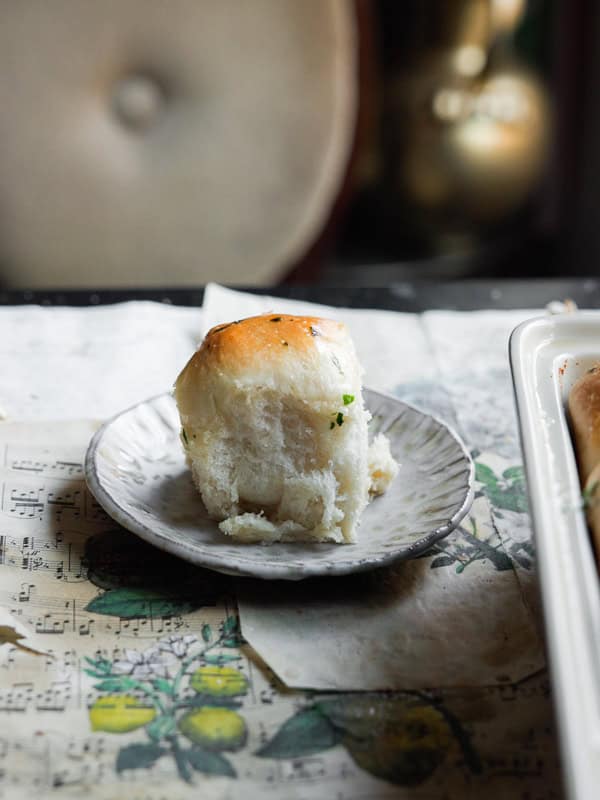 A single soft dinner roll with a golden top sits on a small, textured ceramic plate. The background includes a botanical illustration and a hint of musical notes on paper. The setting is cozy, with muted lighting.